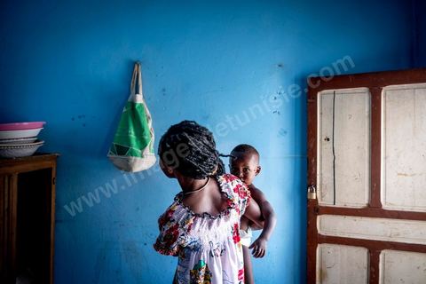 24 juin 2017 : Océane et son fils dans la cuisine. Chez Océane, migrante Ivoirienne à Tunis dans un quartier populaire. Elle vit dans une maison avec 2 chambres qu'elle partage avec son fils Eden et cinq autres ivoiriennes. Océane est arrivée en Tunisie en 2010 comme étudiante et est depuis 5 ans en situation irrégulière, ne lui permettant plus de travailler légalement. Il y a deux ans Eden est né, ce jeune garçon plein de joie est devenu sa raison de vivre, elle souhaite trouver un endroit pour vivre normalement et élever son fils. “J’estime que pour vivre bien Tunisie, il me faudrait entre 800 et 1000 dinars. Généralement, je suis plutôt à 350. Entre les couches et la nourriture, les dépenses sont rapides.” La jeune femme travaille de façon irrégulière : elle a quelques patrons qui l’appellent, selon leur besoin, pour faire le ménage chez eux. Une journée lui rapporte entre 25 et 30 dinars.  