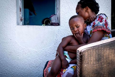 24 juin 2017 : Océane et son fils Eden devant la cuisine qui donne sur la cour. Chez Océane, migrante Ivoirienne à Tunis dans un quartier populaire. Elle vit dans une maison avec 2 chambres qu'elle partage avec son fils Eden et cinq autres ivoiriennes. Océane est arrivée en Tunisie en 2010 comme étudiante et est depuis 5 ans en situation irrégulière, ne lui permettant plus de travailler légalement. Il y a deux ans Eden est né, ce jeune garçon plein de joie est devenu sa raison de vivre, elle souhaite trouver un endroit pour vivre normalement et élever son fils. “J’estime que pour vivre bien Tunisie, il me faudrait entre 800 et 1000 dinars. Généralement, je suis plutôt à 350. Entre les couches et la nourriture, les dépenses sont rapides.” La jeune femme travaille de façon irrégulière : elle a quelques patrons qui l’appellent, selon leur besoin, pour faire le ménage chez eux. Une journée lui rapporte entre 25 et 30 dinars.  