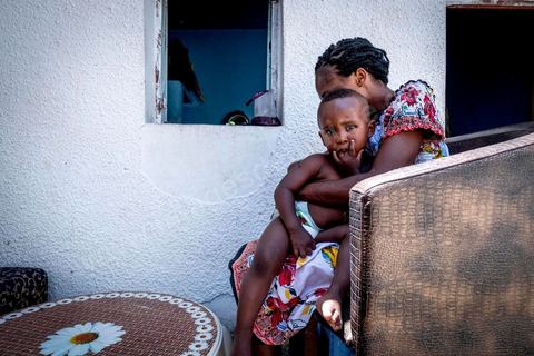 24 juin 2017 : Océane et son fils Eden devant la cuisine qui donne sur la cour. Chez Océane, migrante Ivoirienne à Tunis dans un quartier populaire. Elle vit dans une maison avec 2 chambres qu'elle partage avec son fils Eden et cinq autres ivoiriennes. Océane est arrivée en Tunisie en 2010 comme étudiante et est depuis 5 ans en situation irrégulière, ne lui permettant plus de travailler légalement. Il y a deux ans Eden est né, ce jeune garçon plein de joie est devenu sa raison de vivre, elle souhaite trouver un endroit pour vivre normalement et élever son fils. “J’estime que pour vivre bien Tunisie, il me faudrait entre 800 et 1000 dinars. Généralement, je suis plutôt à 350. Entre les couches et la nourriture, les dépenses sont rapides.” La jeune femme travaille de façon irrégulière : elle a quelques patrons qui l’appellent, selon leur besoin, pour faire le ménage chez eux. Une journée lui rapporte entre 25 et 30 dinars.  