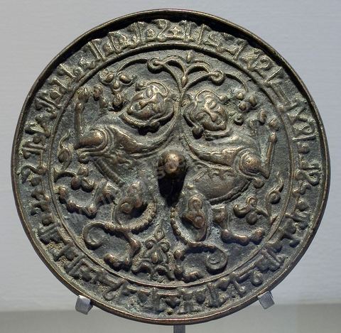 MIROIR EN BRONZE 'AÏNÉ '. Khorassan, 13  siècle.Au centre, décor de chimères adossées : corps de fauve ailé et figure humaine couronnée.Tout autour une inscription bénédictoire en écriture dite 'coufique' : "Gloire et pérennité, succès  et splendeur, élévation et louange, félicité et grandeur, souveraineté et prospérité, puissance et don de Dieu à son possesseur éternellement ".