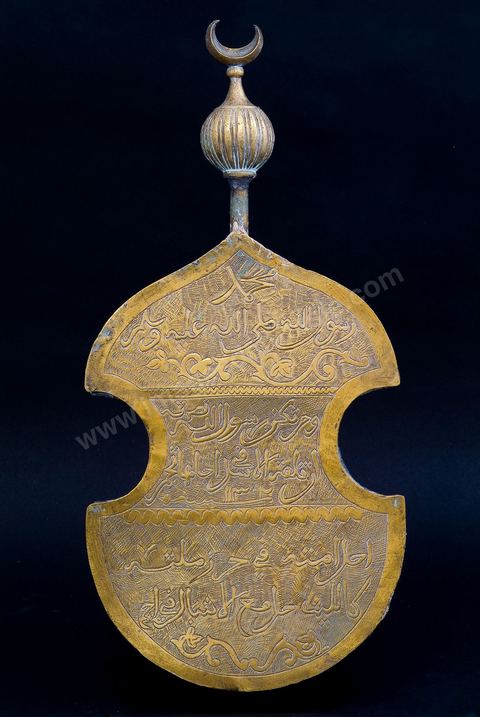 EMBLÈME DE CONFRÉRIE RELIGIEUSE CHADHILIA  OU ISAOUIA. Tunis, 1302H/1884.En cuivre ciselé, gravé d'une inscription. La forme de la  découpe et le couronnement en sphère côtelée et croissant  sont d'inspiration ottomane. Après la prière et le salut du prophète Mohammad, l'inscription reproduit deux célèbres vers extraits de la 'Burda', la Cape du prophète. Un poème à la gloire de Mohammad, composé  par al-Busaïri, chef de la confrérie Chadhilia d'Egypte(13  siècle). "Celui qui, par le prophète est soutenu, est à même d'immobiliser  les lions. Il a placé sa nation sous la protection des siens, tel le lion ses lionceaux". En Tunisie, l'attrait traditionnel qu'exerce la figure souveraine du lion, l'habitude d'agir sous le patronage du  prophète ont conféré à ce poème et plus particulièrement à ce passage une aura singulière. Les beys de la dynastie huseïnide (18 -20  siècle), qui étaient souvent membres ou protecteurs de la confrérie Chadhilia, les faisaient graver sur leur sceaux. Fondée en Tunisie par Abou al Hassan Chadhilî (Maroc 1196/ Egypte 1258), la Chadhilia, est à l'origine une voie d'une grande sobriété qui préconisait le dépouillement de l'âme humaine jusqu'à son extinction en dieu. Sans recourir aux techniques de l'ivresse spirituelle, elle insistait sur le 'resserrement en soi ' et le contrôle de sa 'dilatation'.Dans la pratique, à Tunis où elle est encore suivie, il s'agit  d'une voie médiane, mondaine et spirituelle à la fois, ne reniant pas les agréments de la vie et donnant, de fait, refuge aux pratiques corporelles excentriques de la Isaouia. Les adeptes de la Isaouia, convaincus de leur insensibilité à la douleur, avalent des clous et des bris de verre et se transpercent le corps. La grotte de 'Sidi Bel Hassan Chadhilî ' est encore aujourd'hui l'objet d'une dévotion populaire. Lieu nimbé de mystères et chargé de miracles, il est selon les propres paroles de Chadhilî 'visitée la veille de 