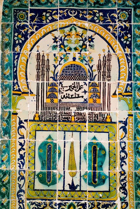 PANNEAU DE CÉRAMIQUE AUX SANDALES DU PROPHÈTE.Tunisie, husseïnide, signé et daté, Khémiri 1216 H./1801Panneau à thème de 'mihrâb', la niche de prière en Islam. Le cadre est orné de rinceaux fleuris de 'lotus' et de feuilles de 'SAZ' ottomanes. Au centre de l'arc, on reconnaît une mosquée à coupole centrale de style ottoman. Six minarets s'élancent, traçant, en graphie coufique à haute hampe, et en miroir, la profession de foi musulmane: Il n'y a de Dieu qu'Allah et Mohammad est son prophète". À leur base se dessine une minuscule porte jaune à claveaux bruns et blancs, donnant une idée de la proportion suggérée. Au centre du mihrâb, de part et d'autre d'une palme, un panneau figurant les sandales du prophète. Au dessous, deux arcs polylobés renfermant une composition florale. La croyance dans les vertus bénéfiques, la baraka, des attributs corporels et vestimentaires du prophète, est encore de nos jours, vivante en Islam. Une sorte de culte est voué à l'empreinte de son pied conservée à Istanbul, et à trois de ses cheveux, enterrés au mausolée de son compagnon, Abou Zamaâ Al Balaoui, à Kairouan, quatrième ville sainte de l'Islam. Du Maroc jusqu'en Inde, les Sandales du prophète ont une place particulièrement éminente, ornant ici les enluminures, là, donnant leur nom à une mosquée. Dans les lieux où elles figurent, elles évoquent la 'visite' ou la 'présence' du prophète. Leur aura assure au lieu protection et patronage. Les cinq motifs en oeilletons, au nombre des doigts de la main, qui ponctuent les sandales, contribuent à la protection et à l'éloignement du mauvais oeil.  La signature de l'artiste, peu courante dans ce genre d'ouvrage et dans l'art  arabe en général, prend, au dessous de la grande coupole, une position centrale, immodeste. Un voeu de piété, le désir d'être enterré auprès du prophète - la mosquée serait celle de son mausolée à Médine -, pourrait la justifier. Protecteur de la demeure 