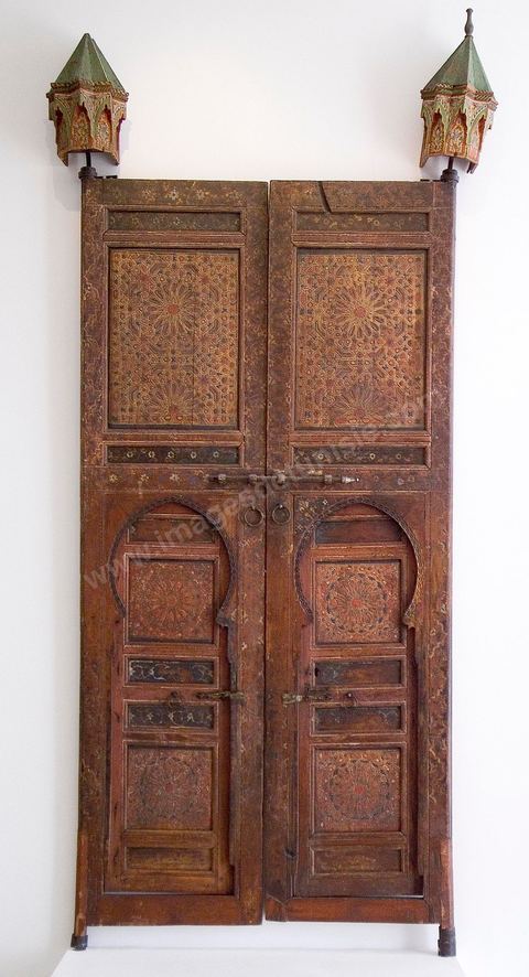 DOUBLE-PORTE À DÉCOR ÉTOILÉ. Maroc, 18   siècle.L'abondance du bois au Maroc, rare dans les pays arabes, a permis la généralisation de son usage en architecture.Toujours ouvragés, souvent peints, poutres, solives, corbeaux, linteaux, piliers, frises et portes se multiplient autant en intérieur qu'en extérieur. Il est dans les pièces, les patios, les façades et dans les passages couverts des ruelles et impasses. Une sculpture particulièrement riche en étoiles et arabesques caractérisaient les mosaïques de bois de l'époque mérinide. La porte exposée ici en hérite la composition étoilée mais réduite à un léger relief. Ses fonds sont décorés de polygones fleuris d'inspiration orientale. D'un effet particulièrement spectaculaire, dans les patios ou les jardins intérieurs des palais et riadh marocains, ce genre de double-porte donne accès à la salle de réception. Elle est souvent couronnée, d'une paire de corbeaux, en pavillon miniature, ornés ici de niches renfermant une fleur d'oeillet. Répondant aux crapaudines inférieurs aménagées dans le marbre, les goujons supérieurs venaient évoluer à l'intérieur de ses corbeaux, préalablement scellés au mur. Durant la saison froide, on se contentait d'ouvrir les portes basses.