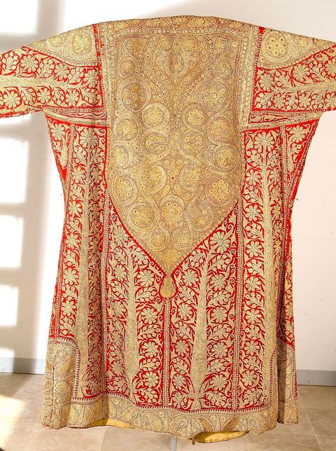 CAFETAN AUX CYPRÈSPerse/Asie Centrale, 19  siècle.Composition très dense caractéristique des costumes d'Asie Centrale. Décors de cyprès et de rinceaux fleuris en fil d'or et d'argent sur fond de soie rouge.Un type de cafetan comparable aurait été porté à Alger durant la période ottomane.