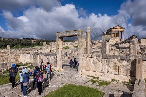 Touristes chinois à dar Lacheb sous fon de temple et capitole. Le site de Dougga, classé au patrimoine mondial de l'UNESCO, Situé sur un éperon dominant la riche vallée de la Mejerdah, le site s'étend sur plusieurs dizaines d'hectares et couvre plusieurs ères historiques. L’excellent état de conservation de la plupart des monuments qui le composent dont certains, tels le capitole ou le théâtre, ont été « remis sur pied » au cours d’une campagne menée au lendemain de la première guerre mondiale par des prisonniers de guerre.