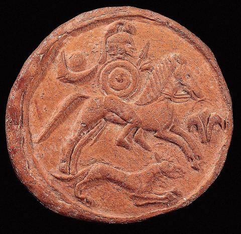 A terracotta medallion depicting a warrior riding bareback on a galloping horse. Wearing a crested helmet and armed with a round shield and a spear, he is accompanied by a dog. To the right of the horse is a lotus or lily flower, and behind the rider are depicted a crescent moon and a sun disk. This may represent a warrior deity (diameter 9 cm, Douimès Necropolis, Carthage, 6th century BCE, Carthage Museum).