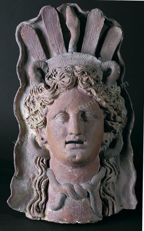 A terracotta mask likely depicting Demeter. The goddess wears a sort of bowl-shaped tiara, framed by a rigid, conch-shaped veil. Vertical, parallel feathers rise from it, with a serpent—a common attribute of chthonic deities—standing out at their center (H. 46 cm, W. 29 cm, Th. 12.5 cm, Salammbô Sanctuary, Carthage, 3rd–2nd century BCE, Carthage Museum).