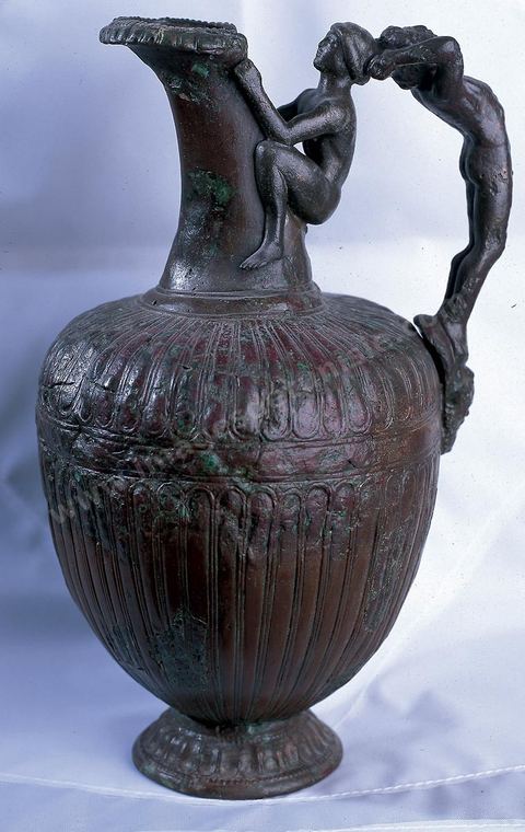 Bronze oenochoe: The body of this bronze oenochoe (wine jug) is adorned with a simple design: fluting on either side of a central band. The handle consists of two figurines: a teenager clinging to the neck of the vessel, which he holds between his legs, and a bearded figure whose body is arched backward, leaning against a mask that serves as the lower attachment, with his hands resting on the teenager’s head. Some scholars attribute this oenochoe to Etruria, others to Tarentum or even Carthage (H. 25 cm, diam. 14 cm, Carthage, 5th century BCE, Carthage Museum).