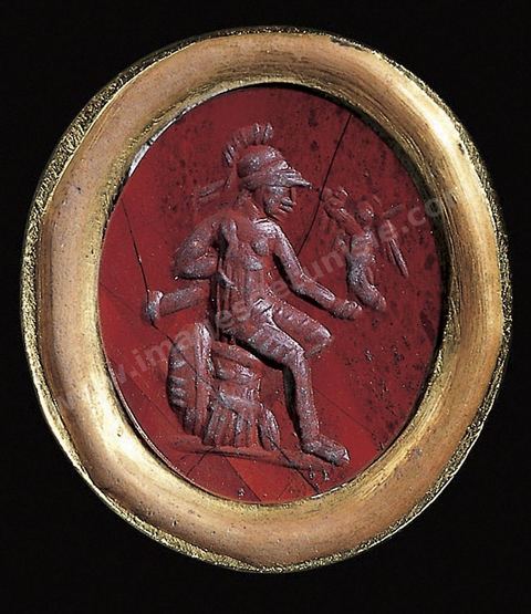 Sur une cornaline cerclée d’or, un personnage assis, casqué et armé, s’apprête à accueillir un personnage qui semble être Amour - section punique - musée du Bardo