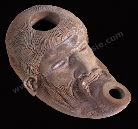 Lampe en terre cuite figurant la tête d’un personnage chevelu, portant barbe et moustache, aux traits finement dessinés et au visage très expressif. Ce type de lampe était, semble-t-il, importé d’Alexandrie ou produit sur place et imité de modèles alexandrins. Celle-ci représenterait une divinité ou un personnage divinisé (Ve au IIIe‑siècles av.‑J.-C., musée du Bardo). Section punique - musée du Bardo